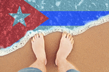 Fototapeta na wymiar Feet on sandy beach with flag Cuba. Top View on sea surf.