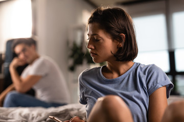 Displeased woman using mobile phone while her boyfriend in sitting in the background.