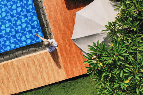 Top view of young unrecognizable woman in white dress and straw hat sitting near swimming pool. without face. Drone photo.Space for text