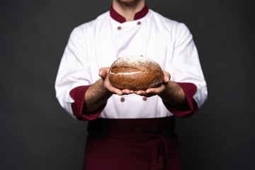 Male baker holding bread loaf and looking at camera isolated on black.