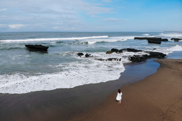 Aerial view of unrecognizable woman in white dress walking near ocean and enjoy summer day on beach. Vacation in Bali. Photo from drone. Copy space. Photo from drone