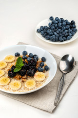 Oatmeal porridge with bananas, walnuts, blackberries, blueberries, honey and mint in a white bowl on a white background. Healthy breakfast and homemade diet food.