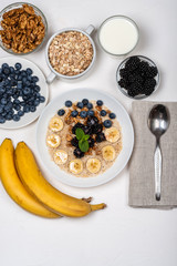 Oatmeal porridge with bananas, walnuts, blackberries, blueberries, honey and mint in a white bowl on a white background. Healthy breakfast and homemade diet food. Top view in flat lay style.
