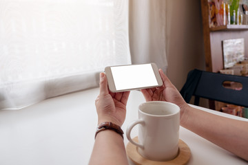 Mockup image of a woman holding white mobile phone with blank desktop screen with a cup of coffee on the table