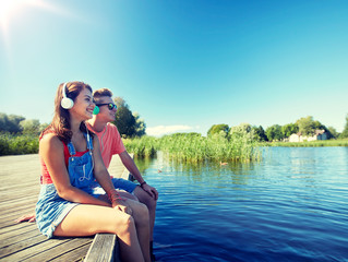 holidays, vacation, love and people concept - happy teenage couple with earphones sitting on river berth and listening to music at summer