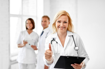 Fototapeta premium healthcare, medicine and profession concept - smiling female middle aged doctor with clipboard and stethoscope showing thumbs up at hospital