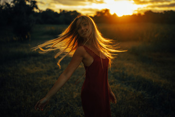 Young blond woman stands on meadow with loose hair lit by sun.