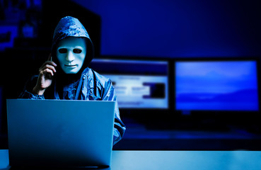 Anonymous computer hacker in white mask and hoodie. Obscured dark face using laptop computer for...