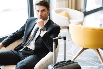 Photo of unshaven young businessman wearing formal black suit sitting in hotel hall with suitcase