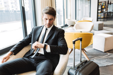 Fototapeta na wymiar Photo of serious young businessman looking at his wristwatch while sitting in hotel hall with suitcase