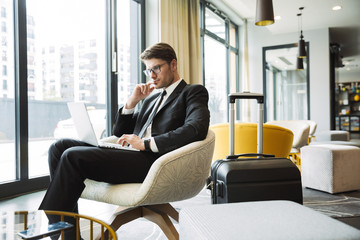 Portrait of beautiful young businessman sitting on armchair with laptop computer and suitcase in...