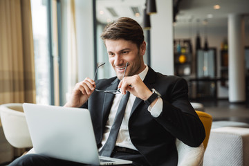 Portrait of smiling young businessman sitting on armchair with laptop computer in hotel hall