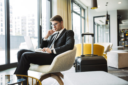 Portrait of beautiful young businessman sitting on armchair with laptop computer and suitcase in hotel hall