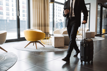 Fototapeta Cropped photo of successful businessman wearing suit holding smartphone and walking with suitcase in hotel lobby obraz