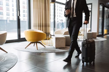 Foto op Plexiglas Bijgesneden foto van succesvolle zakenman in pak met smartphone en wandelend met koffer in hotellobby © Drobot Dean