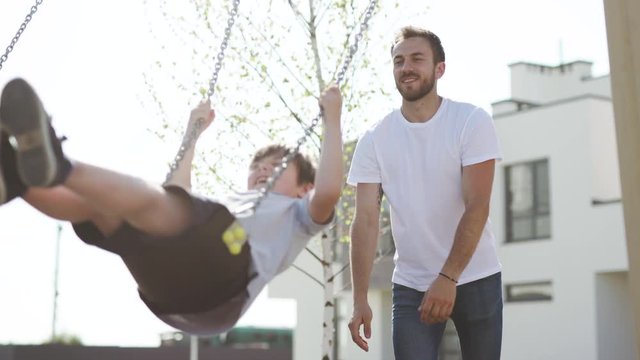 Exciting family time with kids. Cute happy boy swinging and laughing and smiling handsome father standing behind in the playground.