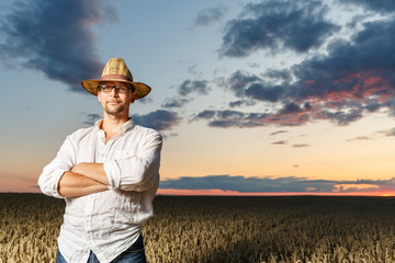 Farmer in a straw hat and glasses standing in a ripe wheat field before sunset.