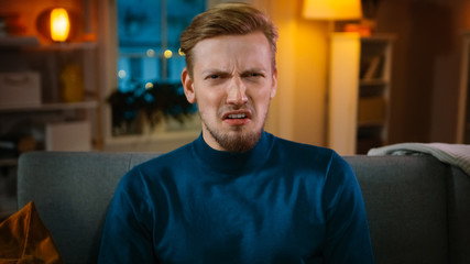 Obraz premium Portrait of a Handsome Man Sitting on a Couch at Home at Night, Watching Gore Horror Movie on TV. He Gets Really Scared, Confused and Disgusted.