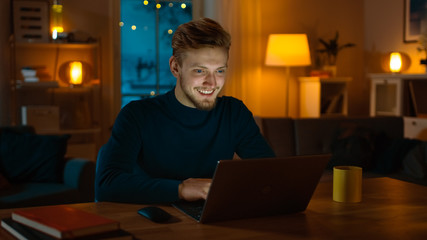 Fototapeta na wymiar Handsome Smiling Man Works on a Laptop Computer while Sitting at His Desk at Home. Cozy Living Room with Warm Evening Lighting Turned on.