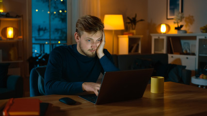 Sleepy and Exhausted Man Works on a Laptop while Sitting at His Desk at Home in the Middle of the Night.