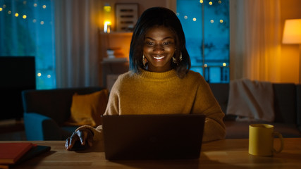 Portrait of Beautiful Smiling Black Girl Using Laptop while Sitting at Her Desk at Home. In the Evening Creative Woman Works on a Computer In Her Cozy Living Room. 