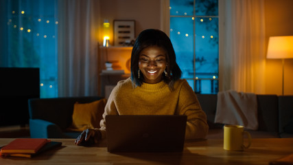 Portrait of Beautiful Smiling Black Girl Working on a Laptop while Sitting at Her Desk at Home. In...