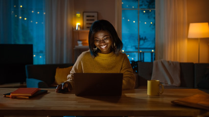 Portrait of Beautiful Smiling Black Girl Working on a Laptop while Sitting at Her Desk at Home. In...