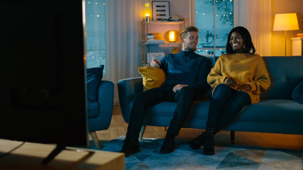 Fototapeta na wymiar Happy Diverse Young Couple Watching Comedy on TV while Sitting on a Couch, they Laugh and Enjoy Show. Handsome Caucasian Boy and Black Girl in Love Spending Time Together in the Cozy Apartment.