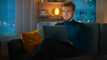 In the Evening Handsome Young Man at Home Sitting on a Couch Works on a Laptop Computer. In the Background Cozy Living Room.