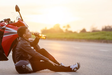 Motorcyclist sitting on the road beside his motorcycle and drinking an alcohol or beer