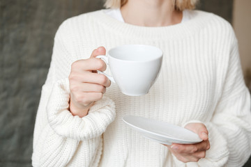 Woman in white knitted sweater holding cup of coffee. Female hands with cup of tea. Close up