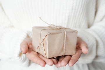 Woman in white knitted sweater giving wrapped gift.  Close up Female hands holding Christmas box present. Christmas, New Year, shopping, preparation on Winter Holidays, donation concept. 