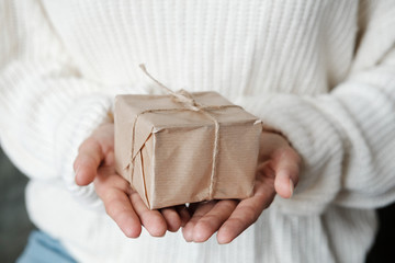 Woman in white knitted sweater giving wrapped gift.  Close up Female hands holding Christmas box present. Christmas, New Year, shopping, preparation on Winter Holidays, donation concept. 