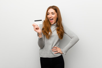 Redhead girl over white wall holding a credit card and surprised