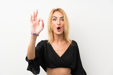 Young blonde woman over isolated white wall surprised and showing ok sign