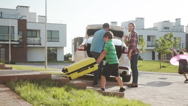 joyful affectionate young family packing suitcases in car trunk near their house. excited girl daughter running of joy in the street wearing pink inflatable lifebuoy. summer trip.