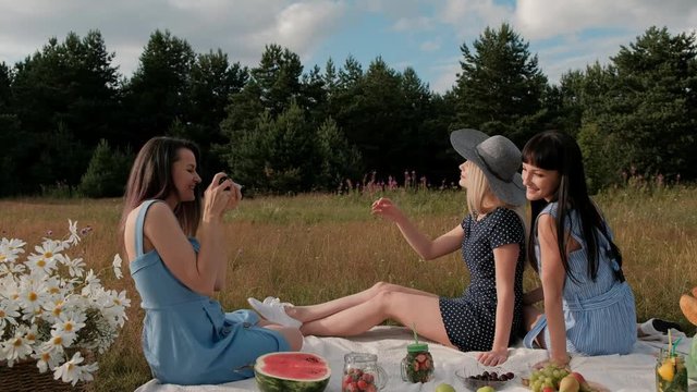 Three young attractive girls on a picnic. The photographer takes pictures on the mirrorless camera of two models. Models pose and watch photos. The concept of outdoor recreation.