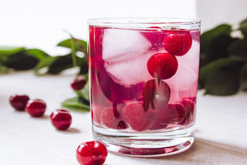 A glass of cherry drink with ice in a glass on a white wooden background. Fresh summer cocktail with cherries and ice cubes.