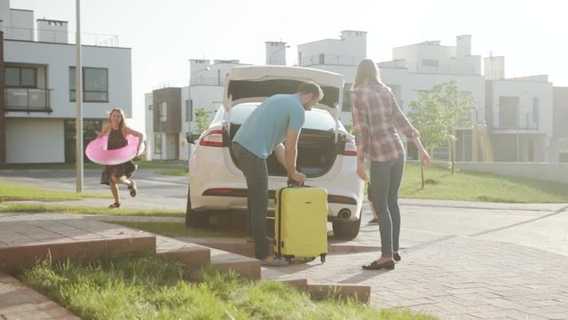 Summer family vacation. Cheerful handsome parents packing and closing up car trunk preparing for a roadtrip. Excited playful kids running after each other playing in the sunny street.