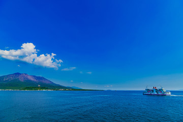 [鹿児島県の観光イメージ] 初夏の快晴の下，噴煙をあげる桜島に向かう桜島フェリーと海上の航跡
