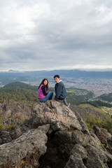 Hispanic couple sitting on top of the mountain turning to see and sniffing with a small town and mountains behind them