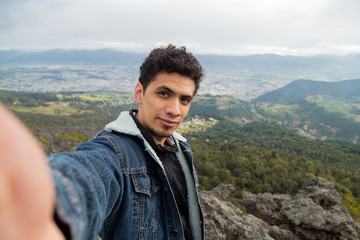 Naklejka premium Handsome Hispanic traveler taking a selfie on top of the mountain with a small town and mountains behind- hiker taking a selfie