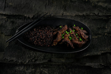 Sticky Asian beef stir fry in salty sweet sauce with black rice in a black clay plate. Directly above top view shot.