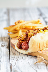Hot dog with chilli, cheddar cheese and mustard. Selective focus with blurred french fries in the background. 