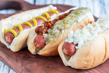 Three varieties of hot dogs over a cutting board. One with coleslaw, one with pickle relish and  chilli and one plain one with mustard only. Selective focus with blurred background. 