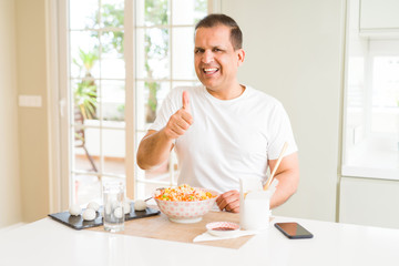 Middle age man eating asian food with chopsticks at home doing happy thumbs up gesture with hand. Approving expression looking at the camera with showing success.