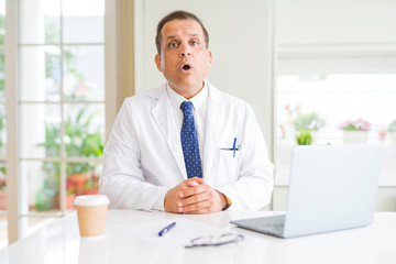 Middle age doctor man wearing white medical coat working with laptop at the clinic afraid and shocked with surprise expression, fear and excited face.