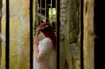 A young woman prays inside a jail ruin. She is  dressed in white and wears a thorn wreath on  her...