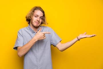 Blonde man over isolated yellow background holding copyspace imaginary on the palm to insert an ad