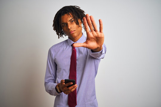 Afro american businessman with dreadlocks using smartphone over isolated white background with open hand doing stop sign with serious and confident expression, defense gesture